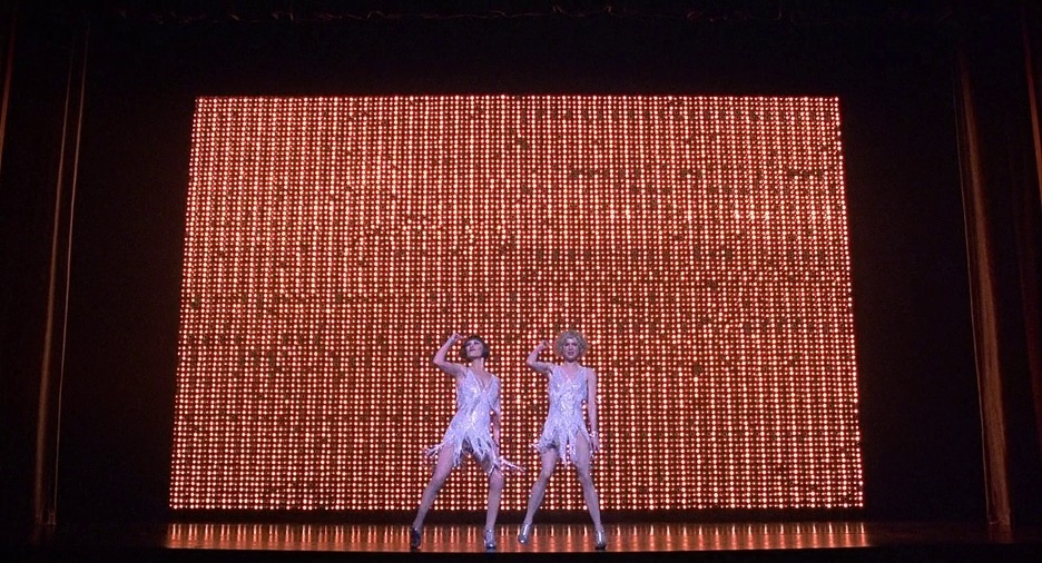 Two women dressed in white flapper dresses dance in front of a red and yellow lit background