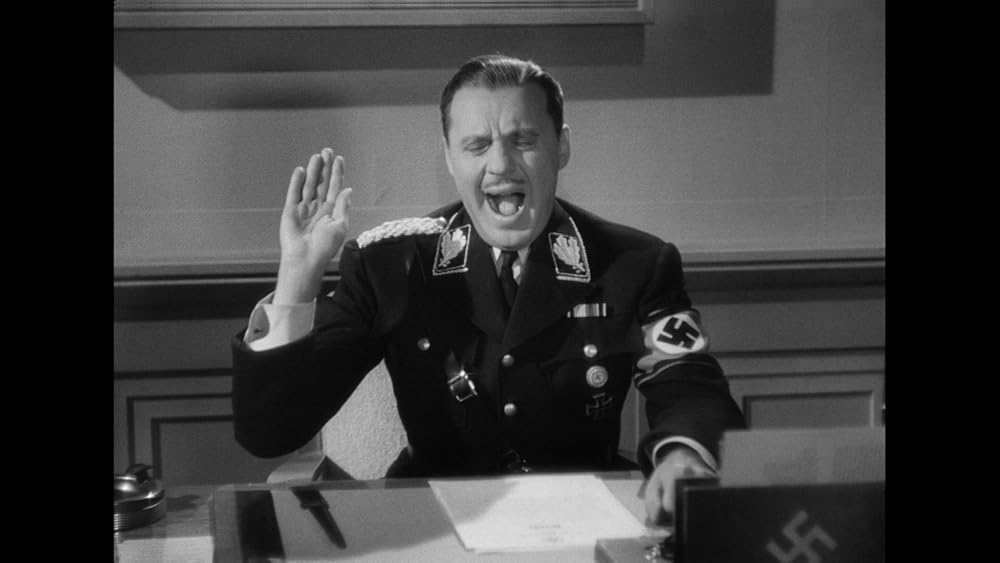 Black and white screenshot of Jack Benny, who sits at a desk in a Nazi uniform with a piece of paper in front of him, and his left arm resting beside it. His right arm is raised in the Nazi salute, and his eyes are closed because he is in the middle of a yawn. He is facing the camera and you only see him from the waist up.