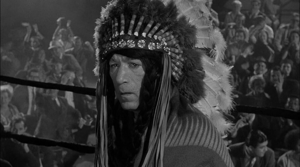 Mountain Rivera, played by Anthony Quinn, stands in a wrestling ring, wearing a Native American headdress, looking pained. The crowd behind him in the smoky arena is loudly booing him.
