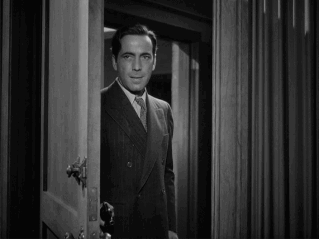 Humphrey Bogart stands behind a partially open door holding a pistol.