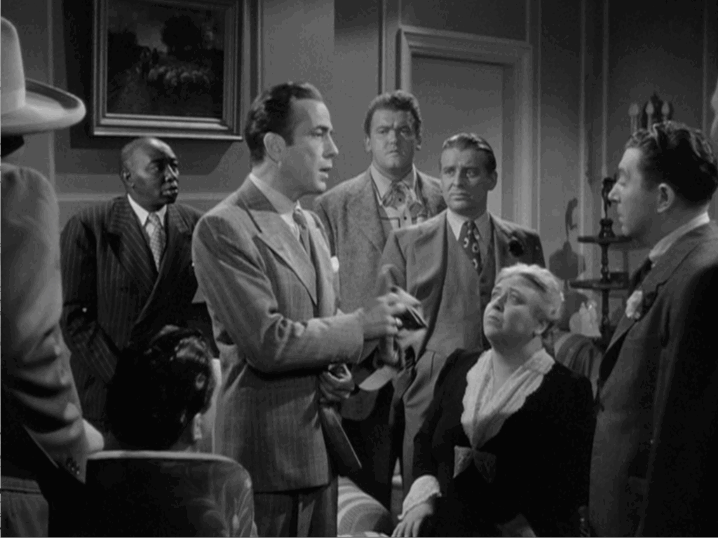 Humphrey Bogart stands in a living room and speaks while a group of people listen to him.