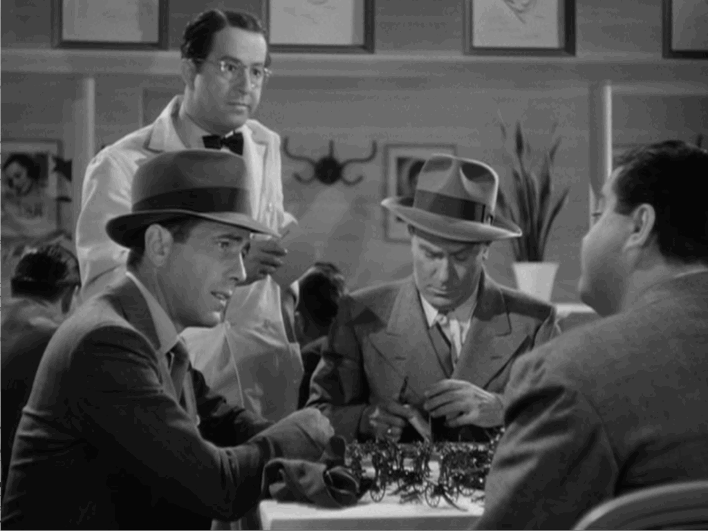 A bespectacled waiter wearing a white suit attends three men sitting at a table covered with children's toy soldiers and cannons.
