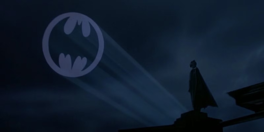 Scene from Tim Burton’s Batman. Batman stands atop a building with the Bat-Signal in the night sky overhead. 