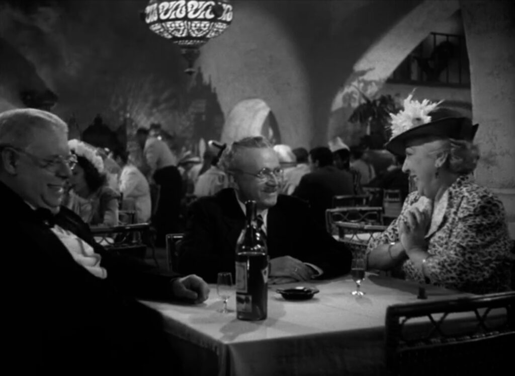 A well-dressed woman and man, both gray-haired, celebrate at a table while the waiter seated with the smiles at the open bottle of cognac.
