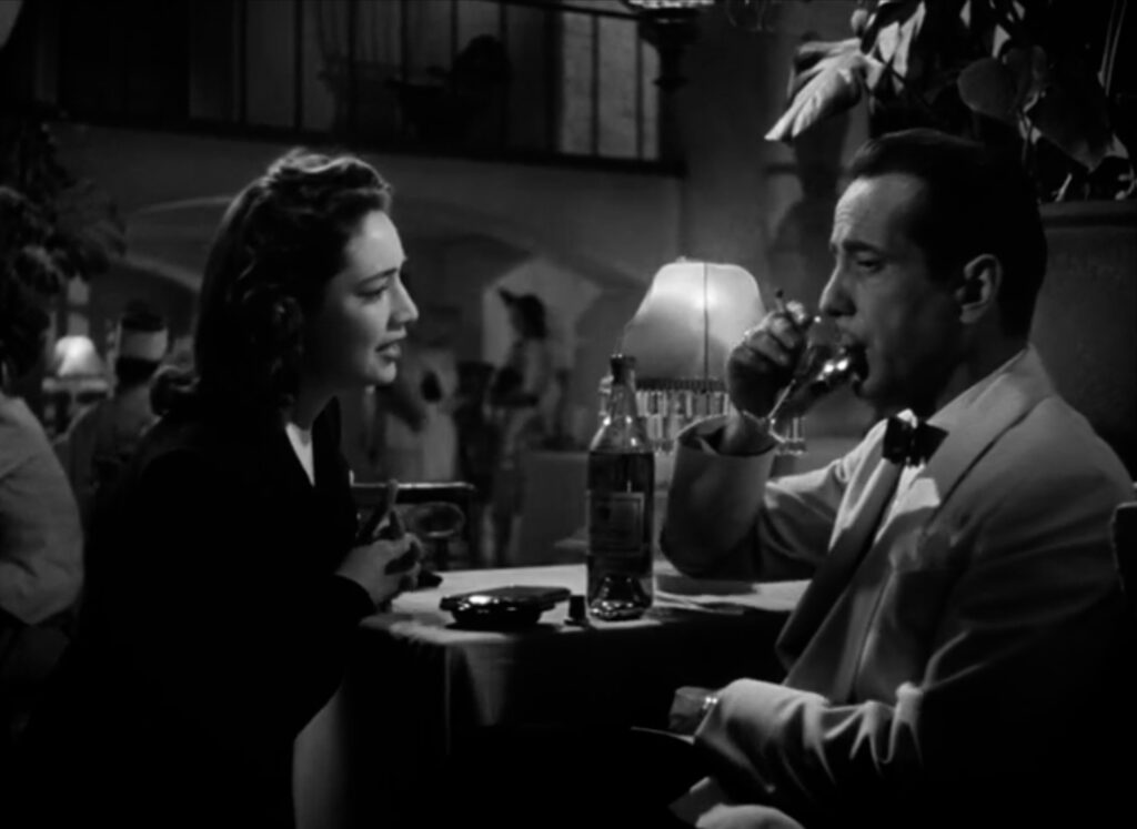 A woman pleading at a cafe table with Rick, who is sipping his cocktail and holding a lit cigarette.