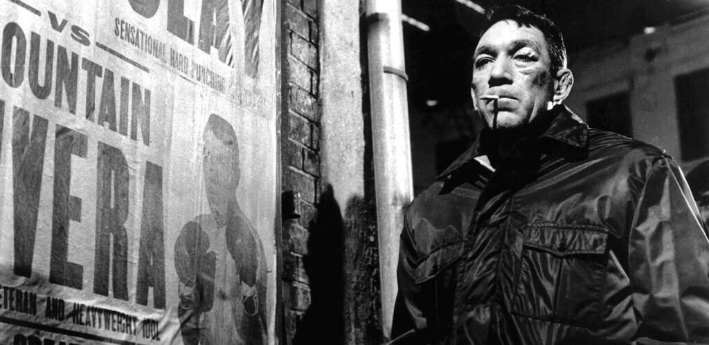 A bruised and bandaged man with a cigarette in his mouth stands next to a boxing poster promoting his own fight. 