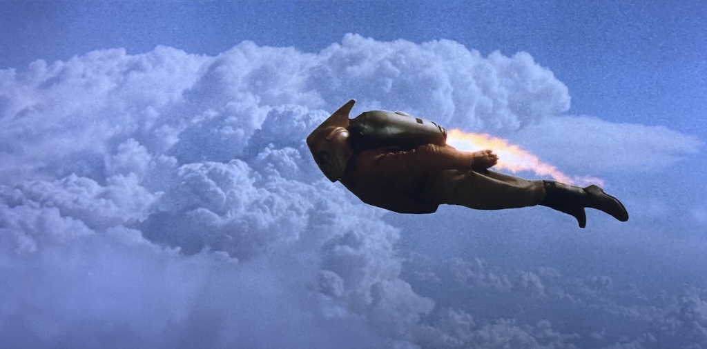 The Rocketeer flies through the clouds in a beautiful sky using his jetpack. 