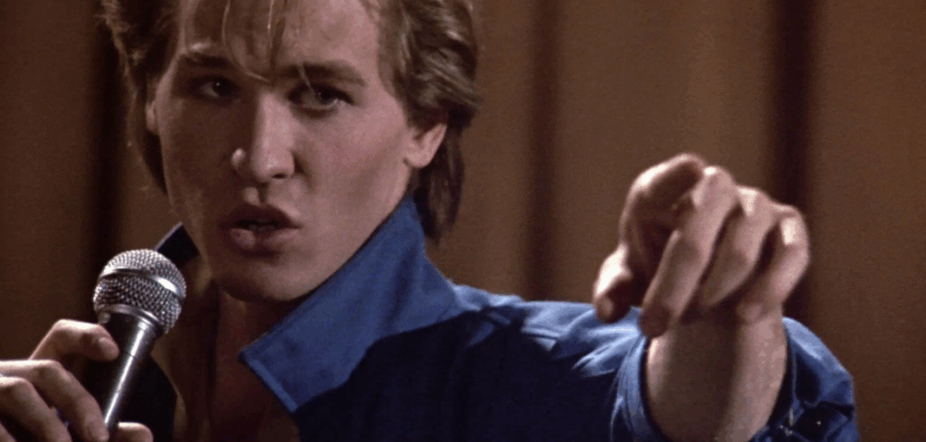 a close up shot of young Val Kilmer earing a blue collared shirt, holding a microphone close to his mouth with his right hand, and pointing with his left hand.
