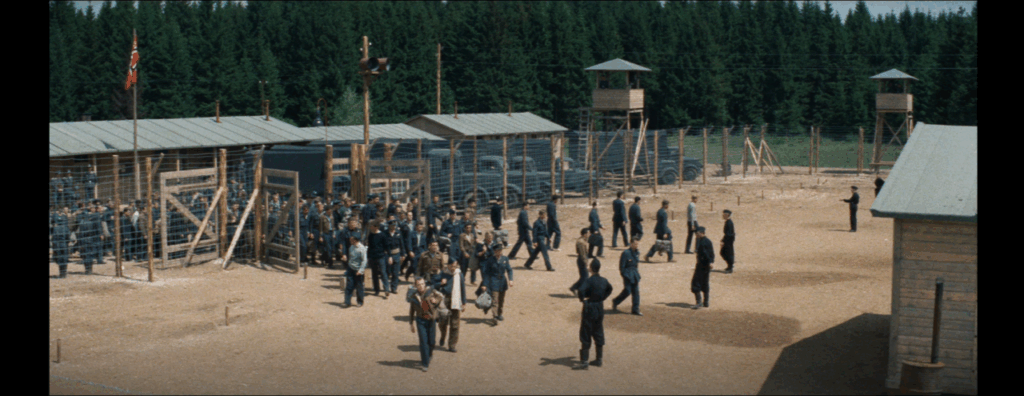 A large group of prisoners walks through the gates and into the Stalag Luft III camp, where German guards await them