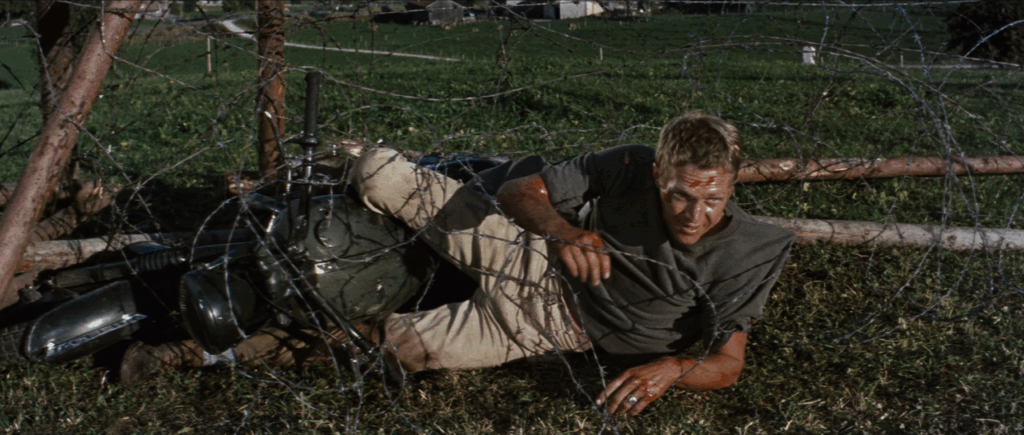 Virgil Hilts (Steve McQueen) covered in blood and tangled in a barbed wire fence and struggling to get out, a motorcycle he was riding lying beside him