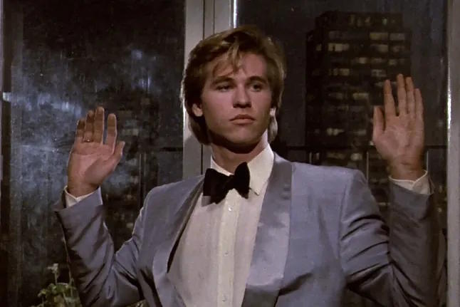Val Kilmer dressed in a baby blue suit coat with his hands up.