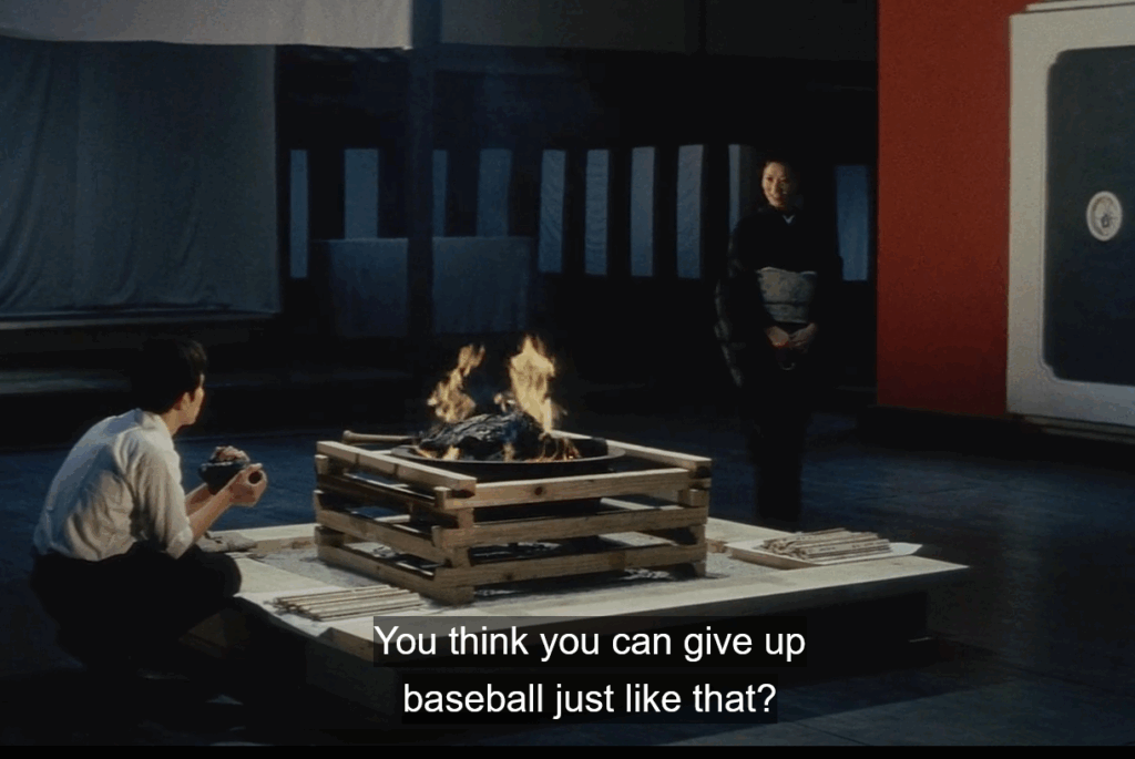 In a dim traditional Japanese interior, a man in a white shirt sits cross-legged beside a small, sacrificial fire. Across from him, a woman in a black kimono with a silver obi stands smiling faintly. The space is lit warmly by the flames, with papers laid around the table. White subtitles read: “You think you can give up baseball just like that?”
