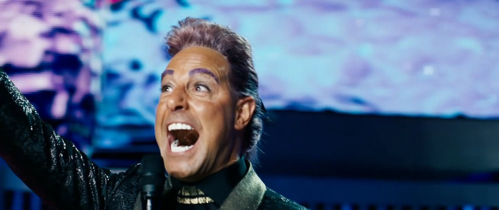 A man with purple hair and eyebrows laughs into a microphone.