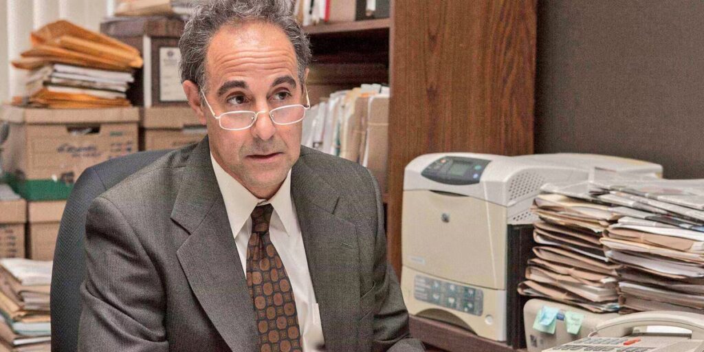 A man in a grey suit and glasses sits in an office full of boxes and files.