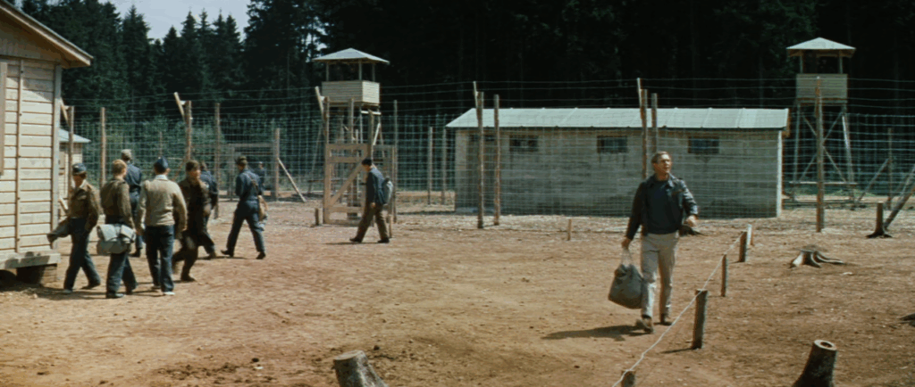 Shot of Steve McQueen in the yard of a prison camp with watchtowers and barbed wire fences in the background
