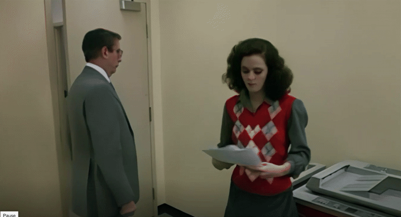 A medium shot of Samantha, dressed in a diamond-plaid red sweater vest with a gray collared shirt and skirt, standing with a stack of fliers at a copier in a nondescript office room. A middle-aged man in wire-rimmed glasses and a drab suit walks by but then turns toward Samantha. The camera cuts to additional fliers falling out of her skirt onto the floor.