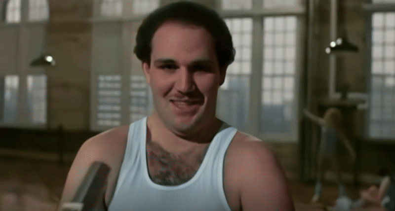 A medium close-up of a hairy man of an uncertain age sporting a white tank-top and a locked-in stare toward the camera featuring a devilish smile, flexing his biceps in cartoonish fashion. An interview mic falls out of frame as he does this at the Heavenly Bodies dance-ercise studio in shallow focus, tall latticed windows and two women stretching in the background. Dark brown hair spills out of his armpits, chest, two-thirds of his head, and upper lip.