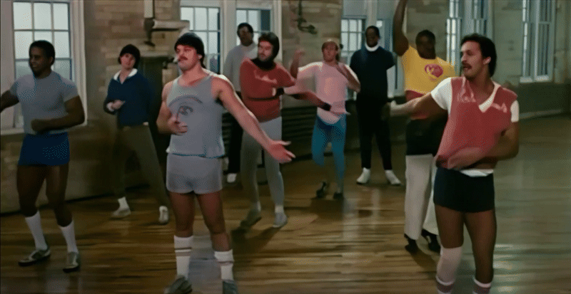 A long shot of nine football players in relatively close proximity to each other dance-ercising with varying levels of conviction. Two men on the right are jumping and extending their arms while a few others are shuffling their feet, but the focal point of the shot is a brawny mustachioed fellow with an emotionless expression moving his arms as limply as possible with zero movement elsewhere in his body. A guy in pink is removing a false breast from under his shirt as he hops in the background too.