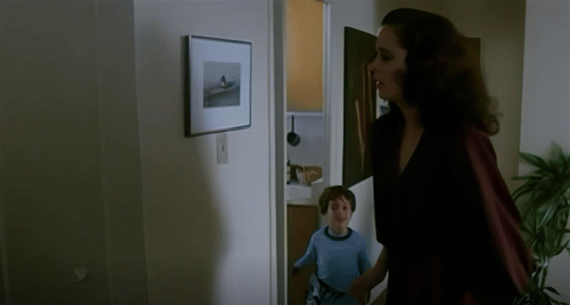A cowboy shot of Samantha in a billowing burgundy suit in her apartment turning toward her young son in pajamas making erratic movements and a silly face in the doorway of their kitchen, disappearing behind the wall, and then Richard Ribiere as Steve suddenly walks out from where the child disappeared. The camera pans right to follow Steve in blue jeans and a beige collared shirt as he smirks at Samantha and walks toward a paper bag of groceries on a table