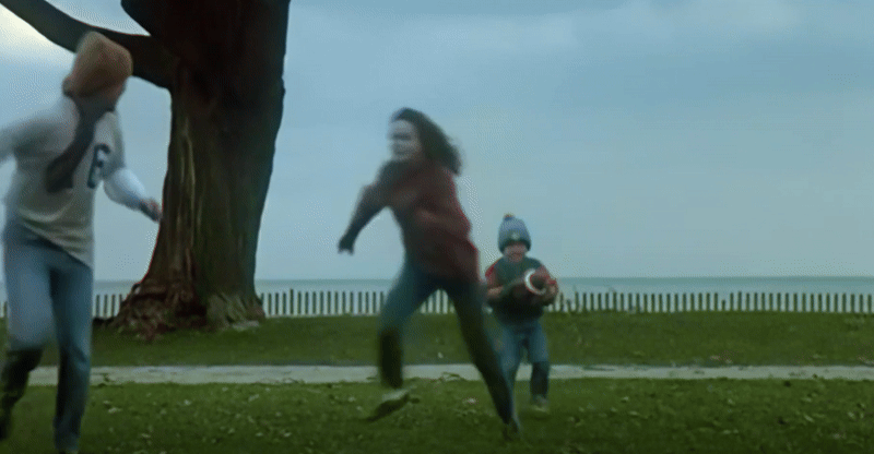 A wide shot of Samantha's small son wearing a winter hat, blue jeans, and coat as he steps backward with a football raised above his head, ready to throw or capitulate to its weight. Samantha and Steve run off to the side and foreground of the frame, ready to receive the ball in the overcast waterfront grass. As they scuttle for a perfect position, gravity does its job and the son falls backward to the ground, suddenly and resolutely.