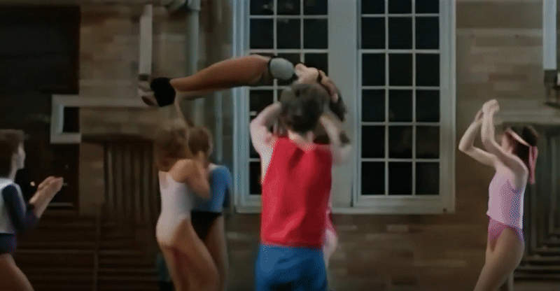A roving long shot of over a dozen of Samantha's students at the dance-ercise studio, three of whom are carrying her face-up in a crucifixion pose in a circle before gently descending her feet-first back to the floor. Clad in a gray and black one-piece with black legwarmers, the crowd around her bounces and claps as their classmates delicately jog their savior around and then down.