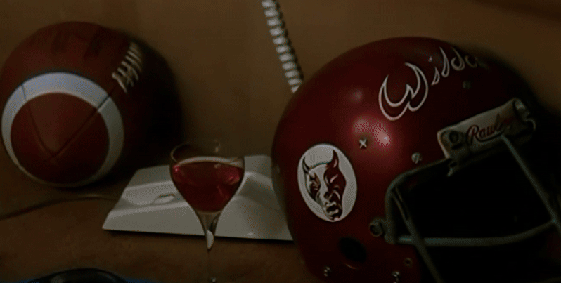 A macro pedestal shot of a football, off-white landline phone base, and red football helmet adorned with the name and image of 'Wildcat', along with a class of rosé in the foreground. As the camera pans up on the khaki-colored sofa, following the curly landline cord to Samantha's face with an arm behind her head, she is revealed to be despondent in her layered white-and-blue top and assorted throw pillows in the background.