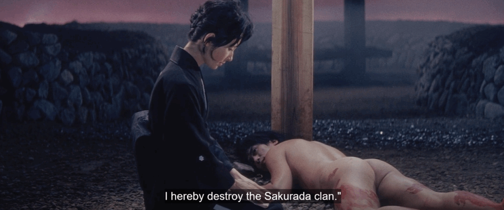 At night in a surreal outdoor setting, a woman in a black kimono kneels beside a bloodied, naked man lying face-down near a wooden post. Low stone walls and a purple sky form the background. Subtitles read: “I hereby destroy the Sakurada clan.”