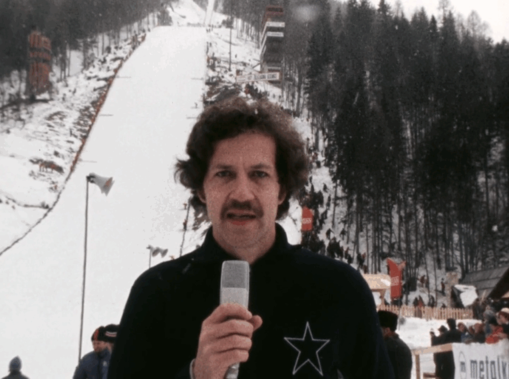 Werner Herzog stands holding a microphone in front of a ski slope