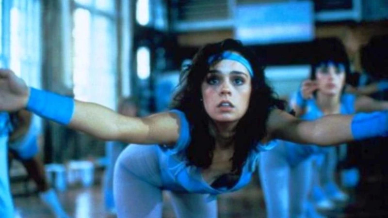 Cynthia Dale as Samantha Blair is front and center in the frame in shallow focus. Brunette and sweaty, adorned by a periwinkle spandex suit along with armbands and headband, she is bending forward with arms extended, seemingly leading a dance-ercise class in a studio with students and light shining through a window behind her. Her facial expression is one of determination and desperation.