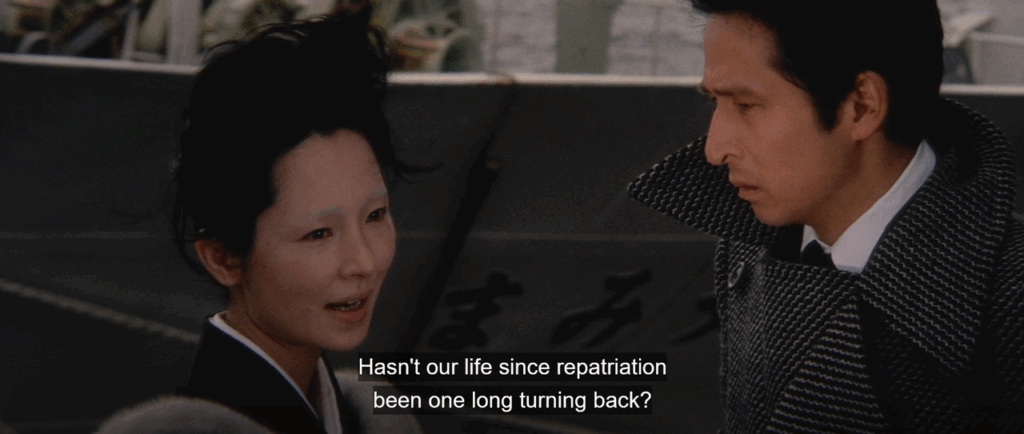A pale-faced woman with faint eyebrows, wearing a black kimono, speaks to a man in a houndstooth overcoat. They stand outdoors near a dock. Subtitles read: “Hasn’t our life since repatriation been one long turning back?"