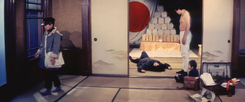 The living room of a Japanese home, where a boy dressed for school stands on the far left, while a smaller child and a man stand on the right. Near the center of the image is a woman lying on the ground.