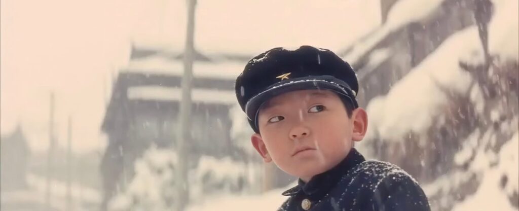 A Japanese boy stands in the snow, wearing a school uniform and a hat.
