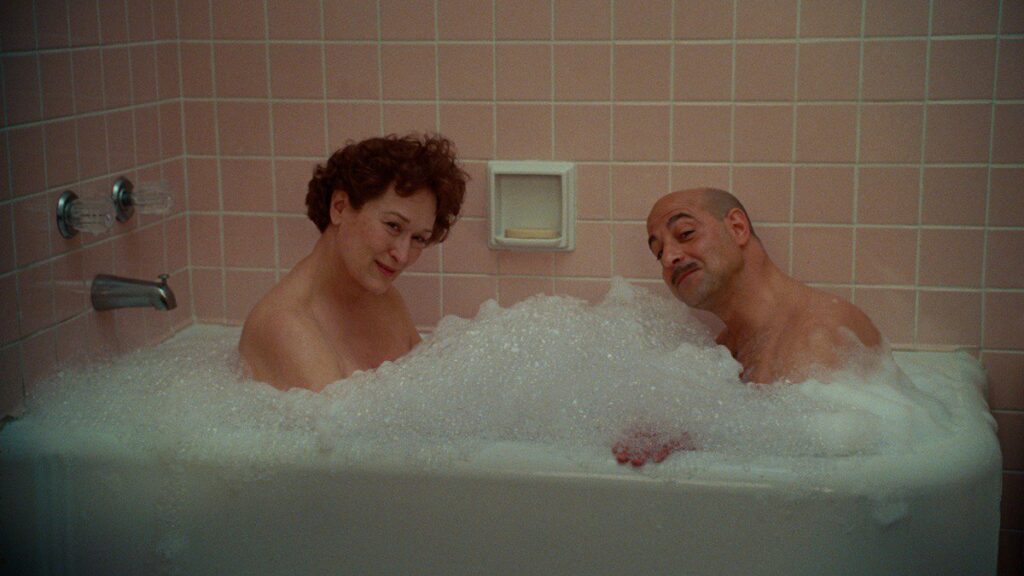 A woman and a man sit in a bathtub full of bubbles and smile at the camera