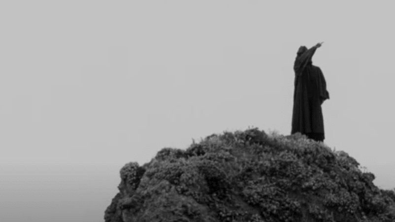 A robed figure stands alone on a hill, holding one hand in the air.
