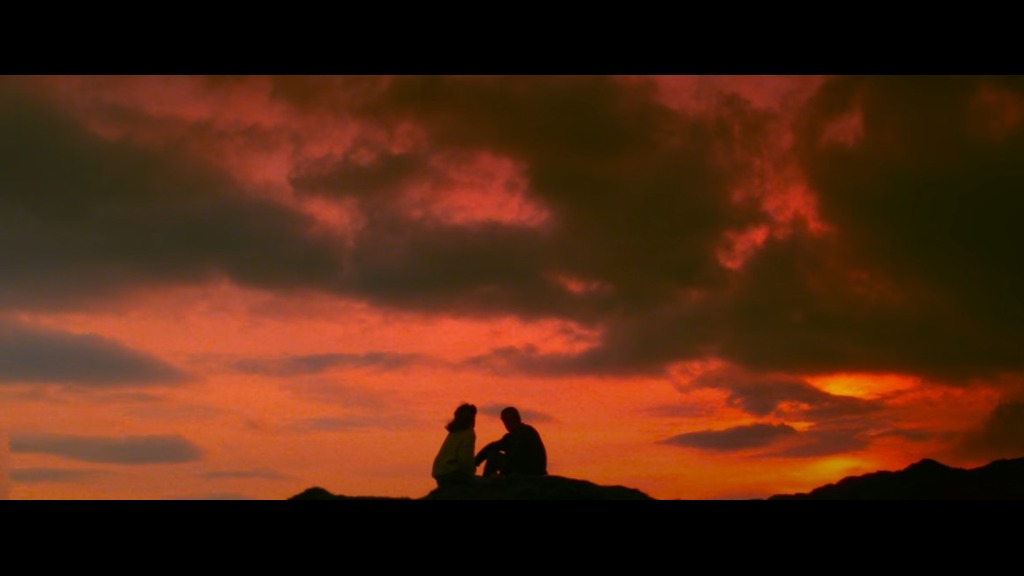 Two figures sit silhouetted against a sunset on a hill