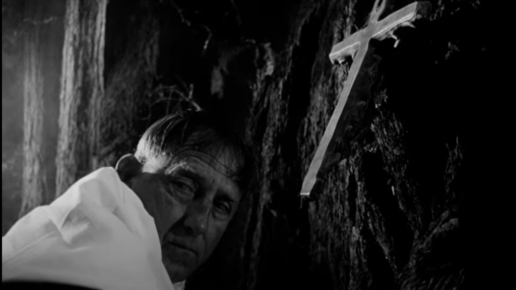A priest sits in front of a tree, where he has hung a silver crucifix facing backwards.