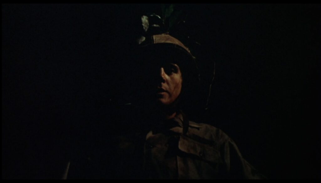 A white male in a military uniform faces the viewer in the dark. Half of his face is in darkness and he looks scared