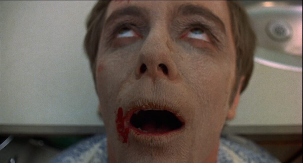 A white male with a grey and aged complexion has his head rolled back on a countertop, his eyes are rolled back in his head in ecstasy. There is a drop of blood on the right side of his mouth