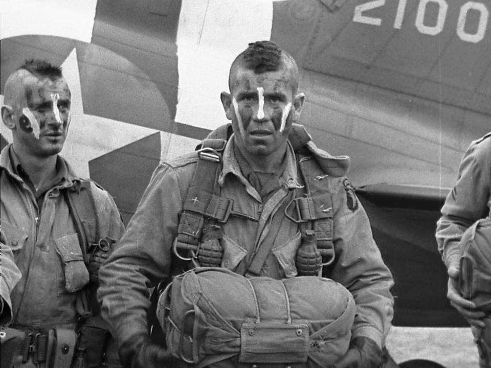 A black and white image of two men standing in front of an aircraft. They are dressed in military fatigues and have streaks of face paint on their nose and cheeks. Both are wearing the Choctaw style of Mohawk.