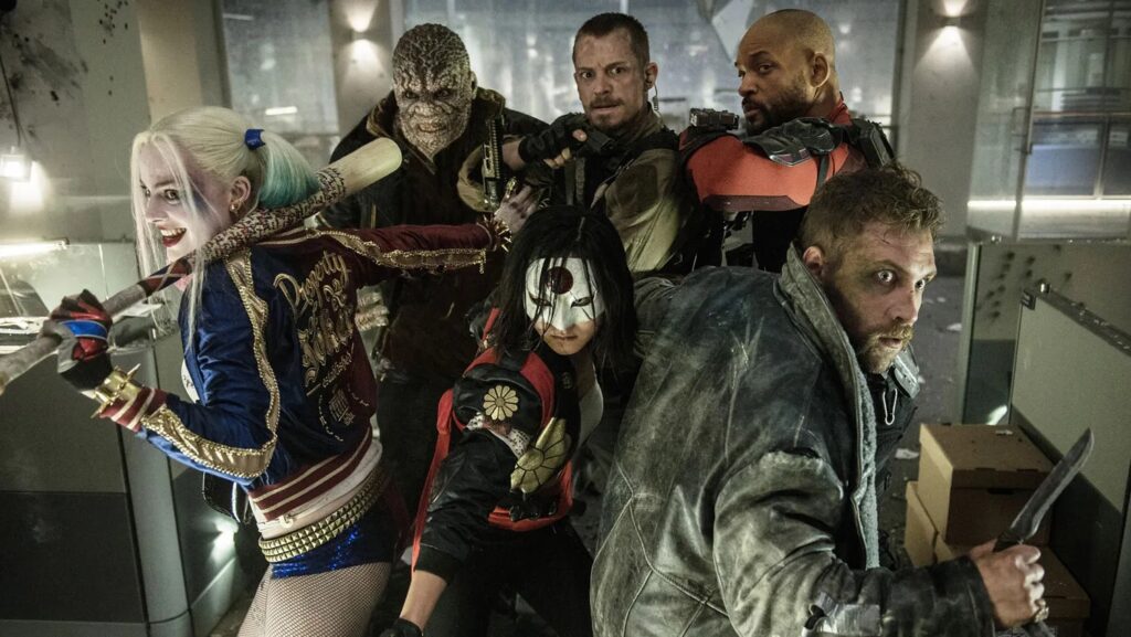 Six garishly dressed super heroes are standing together in an office setting. Top row: Killer Croc, Rick Flag, Deadshot. Bottom row: Harley Quinn, Katana, Captain Boomerang.