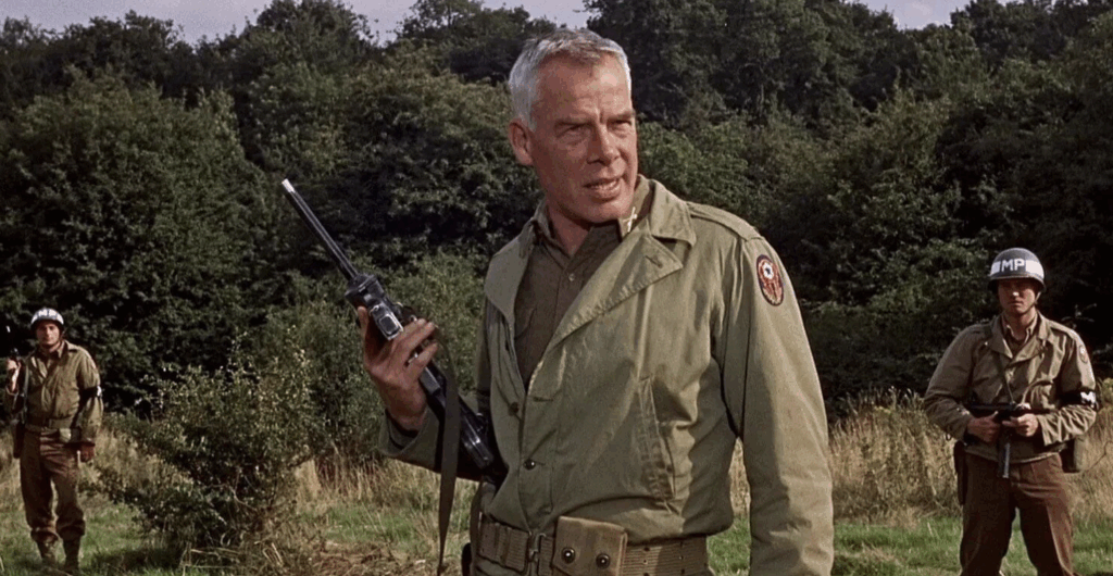 Lee Marvin as John Reisman, standing in the center of the frame. He and two MPs are standing in a field. They are all dressed in green and khaki military fatigues and holding guns.