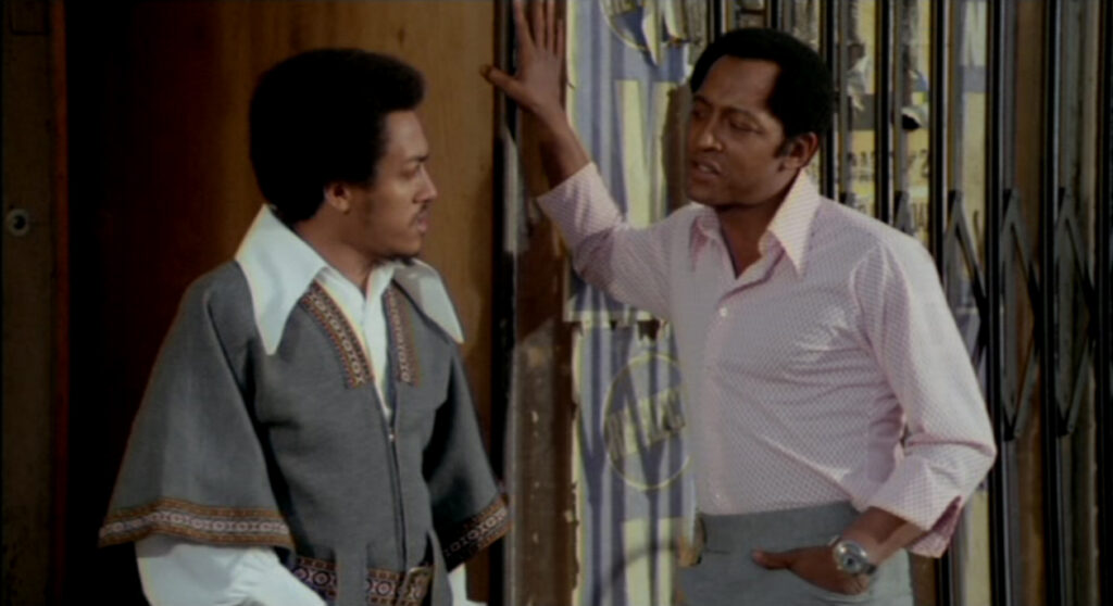  two-shot of Freeman on the right and Shorty (played by Anthony Ray) on the left, standing outside a gated storefront. Freeman is dressed in a pink collared shirt, leaning with his hand up against the doorway, looking down at Shorty with judgment. Shorty's hair is bigger than Freeman's and he is dressed in a more casual white collared shirt covered with a gray patterned African garment as he looks at Freeman with suspicion.