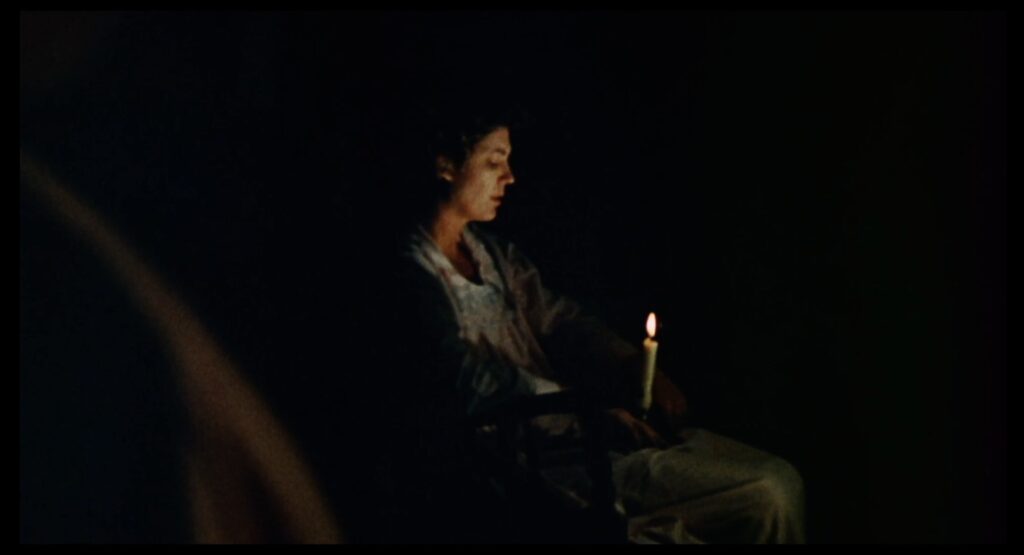 A white woman sits in a chair in the dark, looking mournfully at a lit candle, which illuminates her face