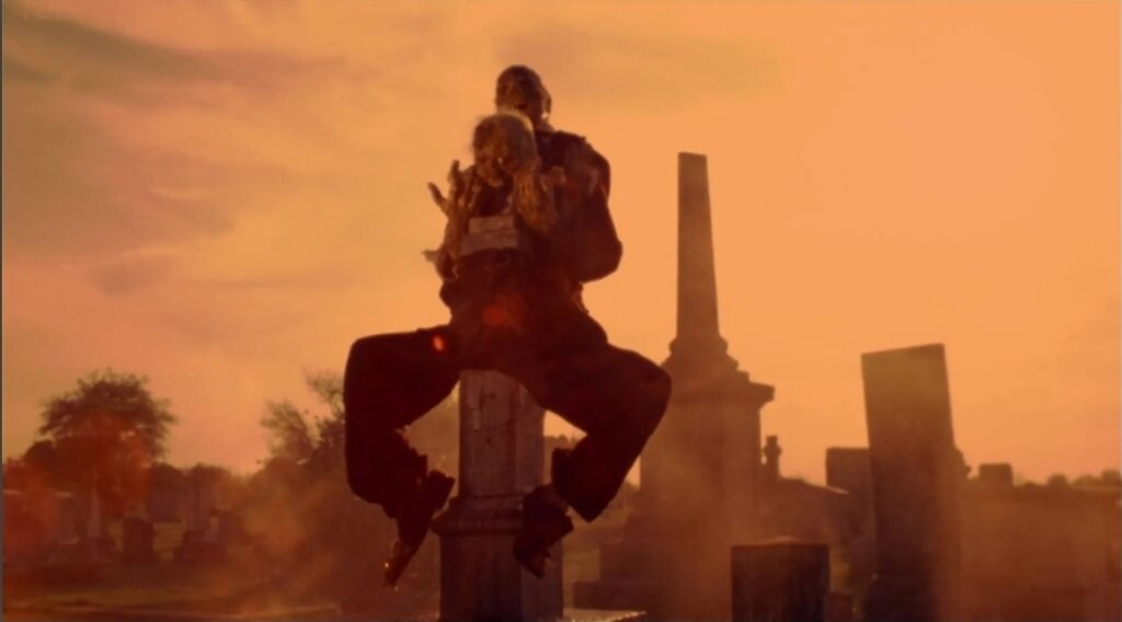 An utterly gruesome pair of fetid dead bodies lashed together and perched on a tombstone, in the arid Texas sky