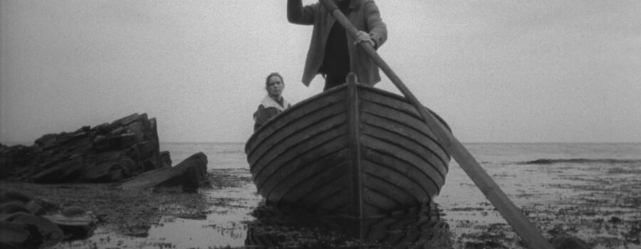 Max Von Sydow (Johan) and Liv Ullman (Alma) arrive at their island getaway. Johan Stands in a large row boat, reflected in dark water, rocky shore nearby.