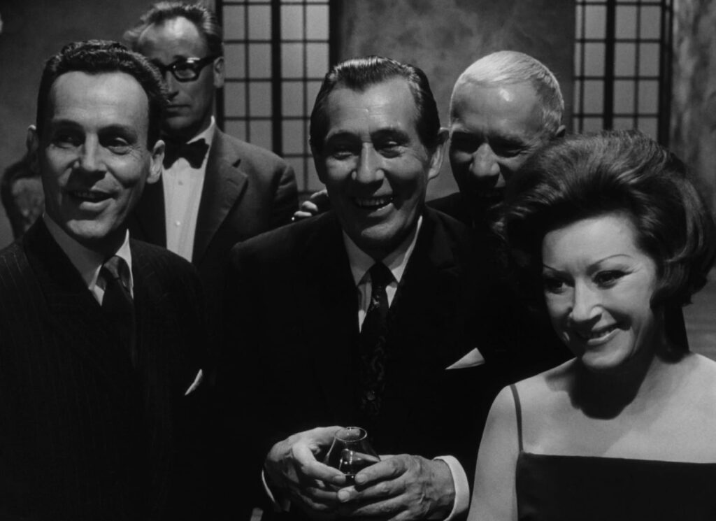 A group of foreboding,  ghoulish dinner guests stand together, laughing and jeering towards an unseen subject in the foreground.