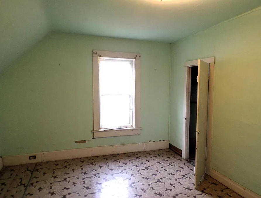 A bedroom on the second floor of a farmhouse, with cracking, lime-green paint, black and white linoleum floors, and a slanted ceiling
