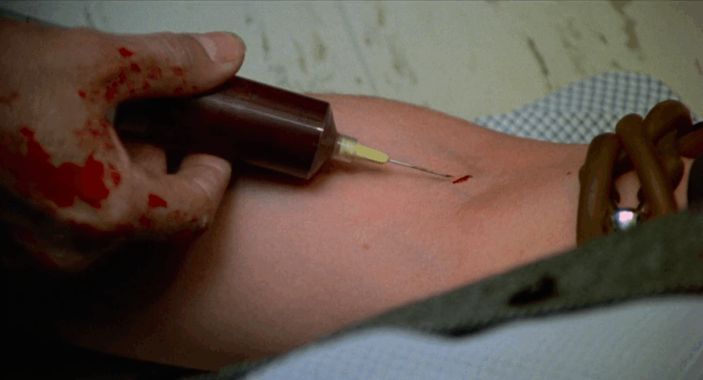  an arm is having blood drawn from it with a large needle. The hand
hodling the needle is covered in blood and looks gray and unhealthy.