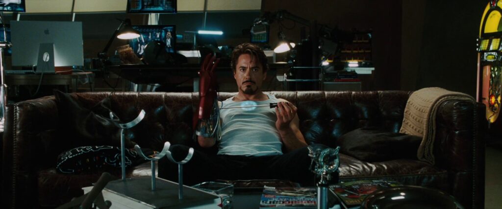 A man wearing a white tank top sits on a couch. He holds a screwdriver up to a mechanical glove on his arm
