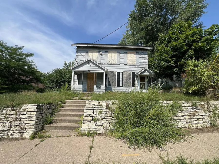 A two-story home with boards over the windows and an uncut lawn.